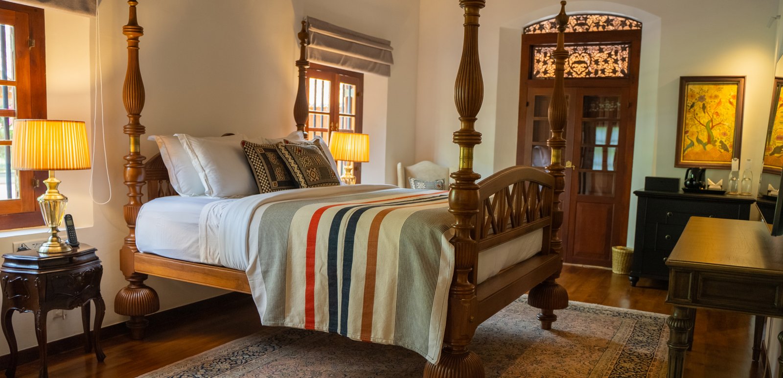 A bedroom with a carved four-poster bed dressed in striped linen, a writing desk and lamps
