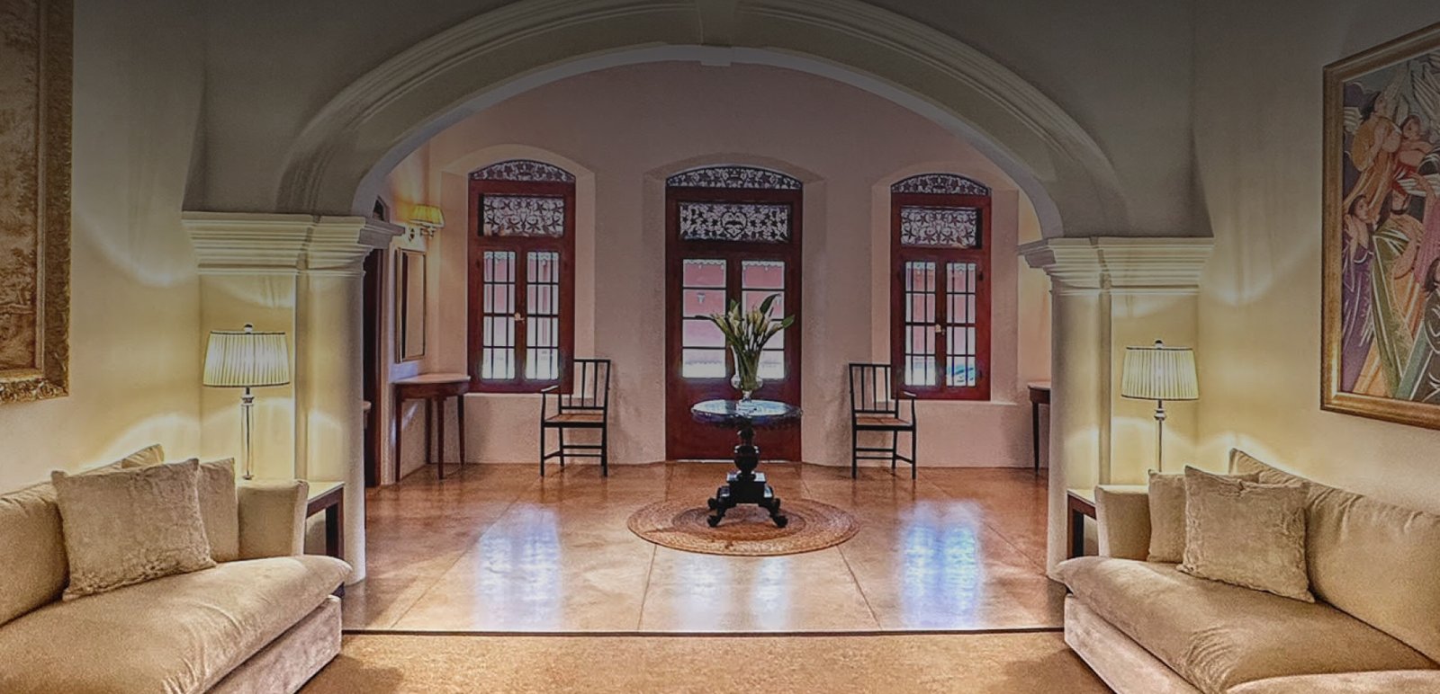 The central hall of the manor under a wide arch, with sofas, a round table and tall louvred windows